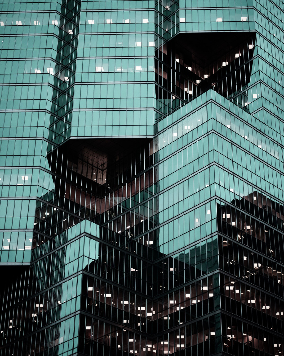 Close-up view of a modern glass office building with geometric shapes, teal-tinted windows, and irregularly stacked sections—an architectural reflection of dynamic businesses adapting to the evolving corporate tax rate. Some windows reveal illuminated office spaces.