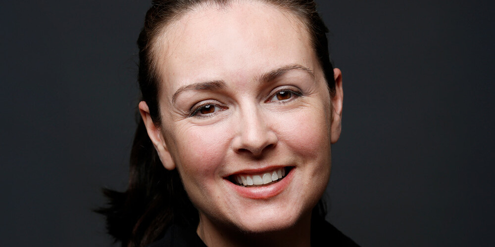 A woman with dark hair pulled back, wearing a black top, smiles warmly at the camera against a plain dark background—an image representing the dedicated team behind About The Canadian Tax Observatory.