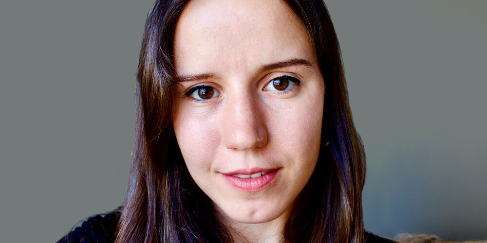 A young woman with long brown hair and brown eyes looks directly at the camera, smiling slightly. She is wearing a black top, with a plain gray background—perfect for an About The Canadian Tax Observatory profile photo.