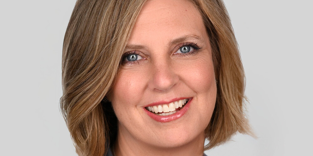 A woman with short, light brown hair smiling, wearing a grey blazer and a silver necklace, posing against a plain light grey background—perfect for an 