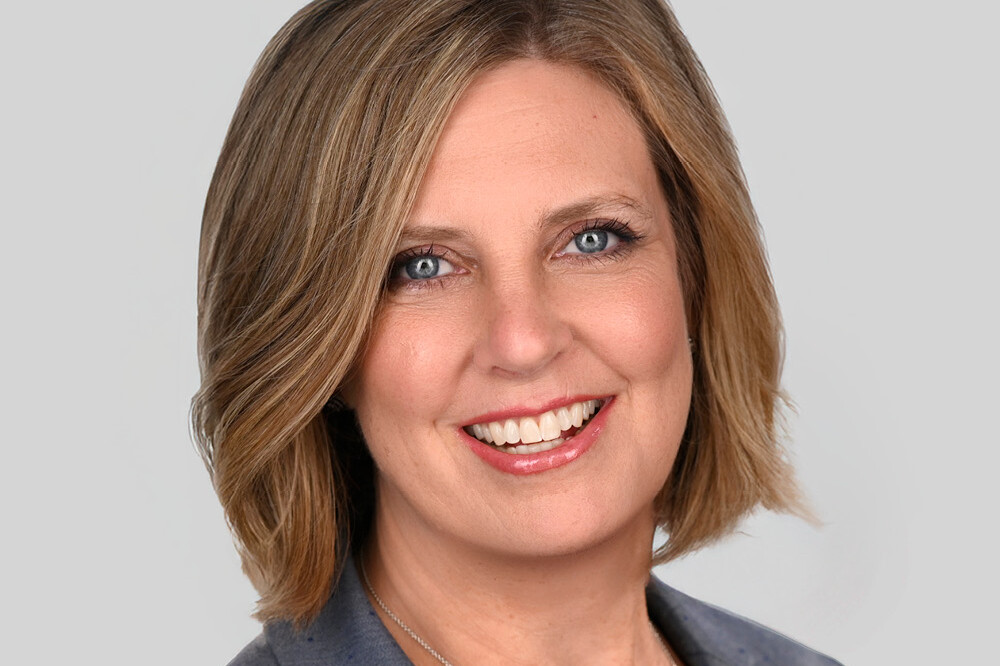 A woman with short, light brown hair smiling, wearing a grey blazer and a silver necklace, posing against a plain light grey background—perfect for an 