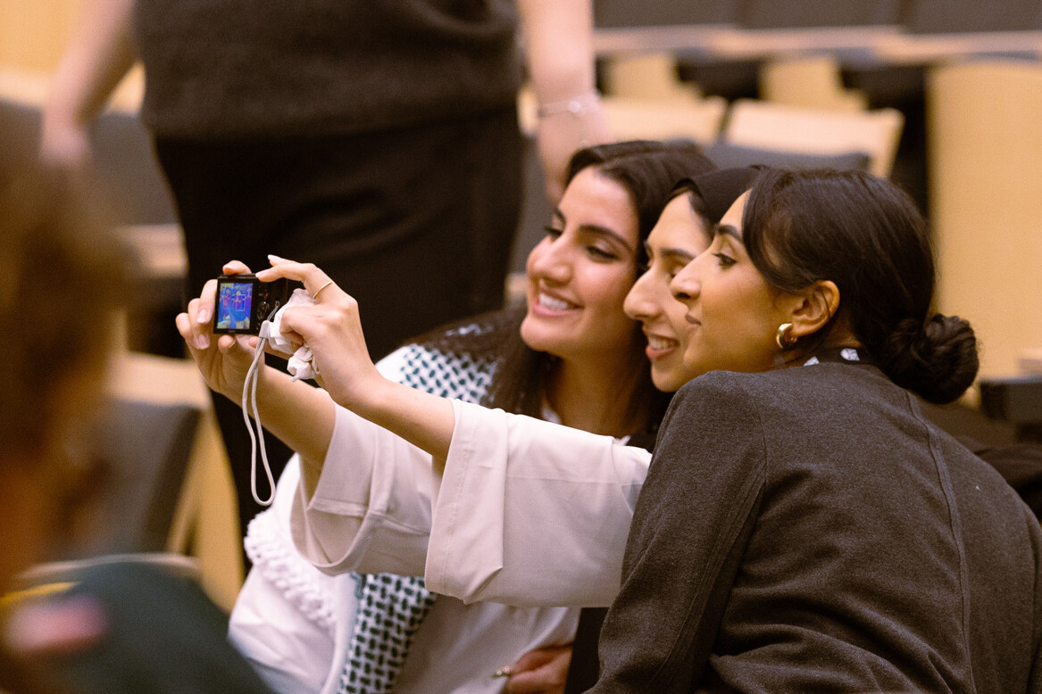 Three women sitting in an auditorium take a selfie together, smiling and posing with a digital camera. The background shows empty wooden seats, hinting at a recent Vancouver public transit funding tax hike contest event. A fourth person is blurred in the foreground.