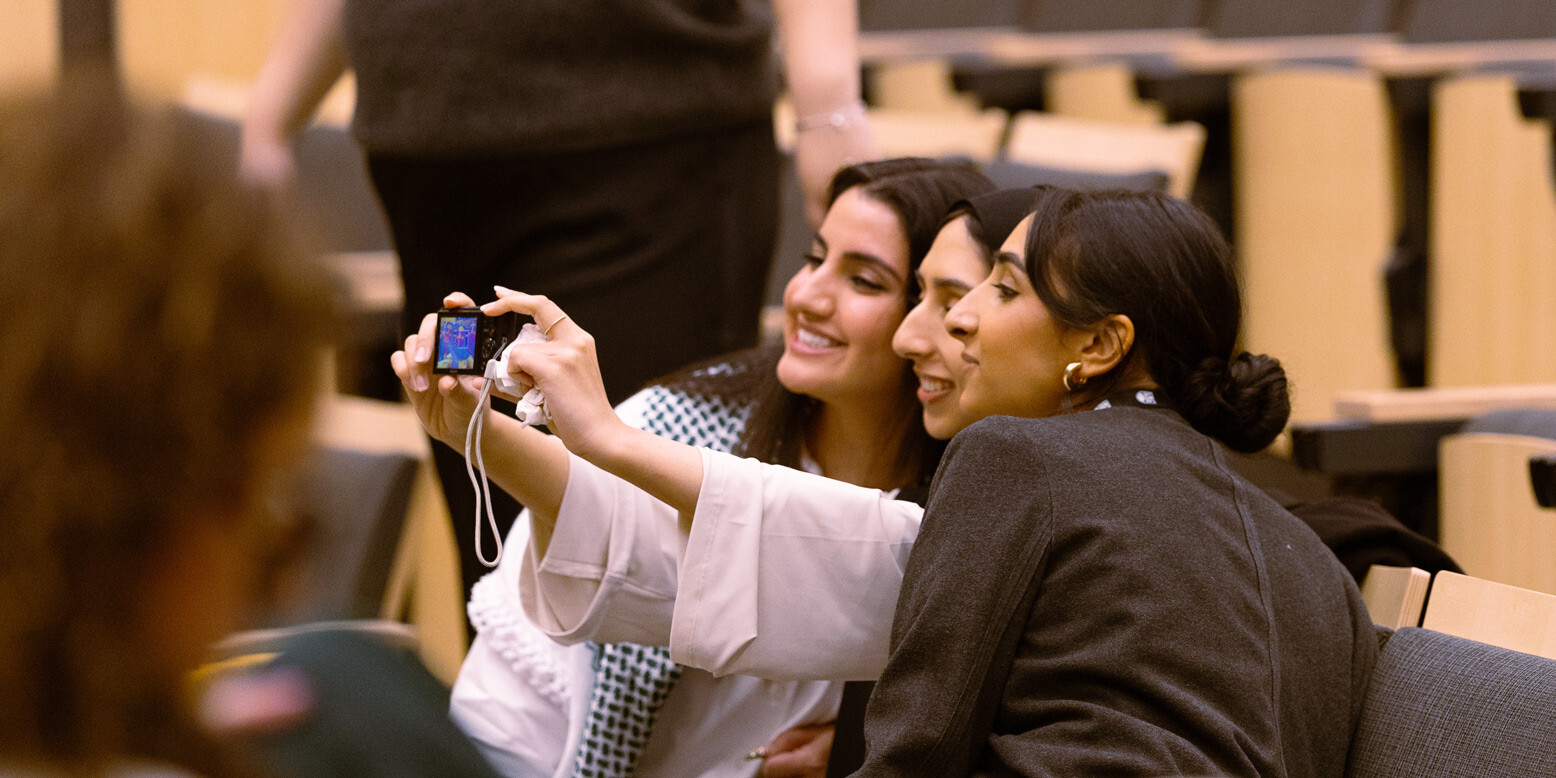 Three women sitting in an auditorium take a selfie together, smiling and posing with a digital camera. The background shows empty wooden seats, hinting at a recent Vancouver public transit funding tax hike contest event. A fourth person is blurred in the foreground.