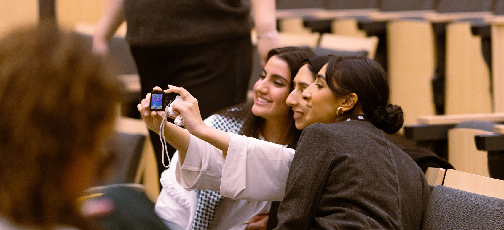 Three women sitting in an auditorium take a selfie together, smiling and posing with a digital camera. The background shows empty wooden seats, hinting at a recent Vancouver public transit funding tax hike contest event. A fourth person is blurred in the foreground.