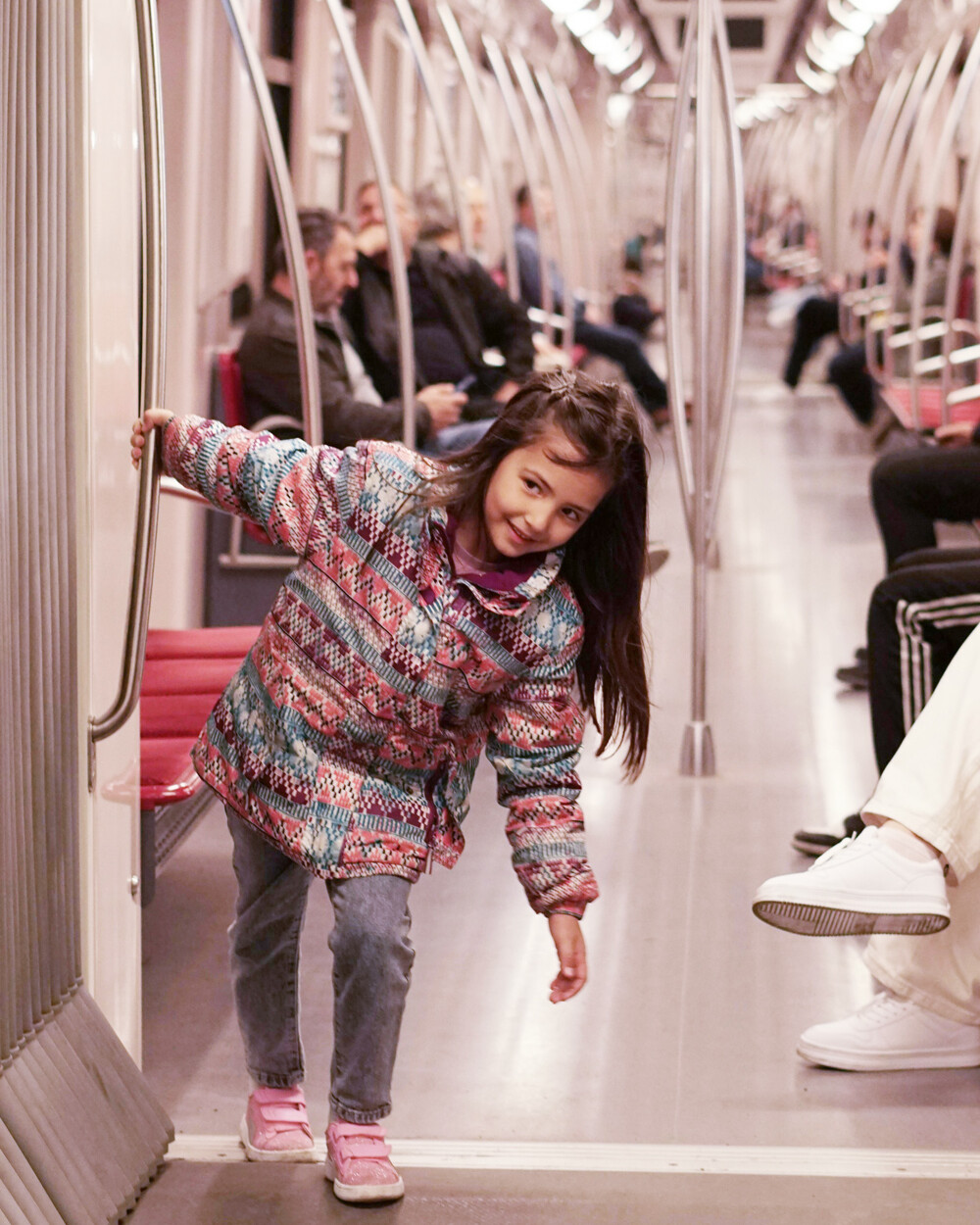A young girl in a colorful coat and pink shoes leans playfully against a pole inside a Vancouver public transit train, while passengers sit along the sides in the background.