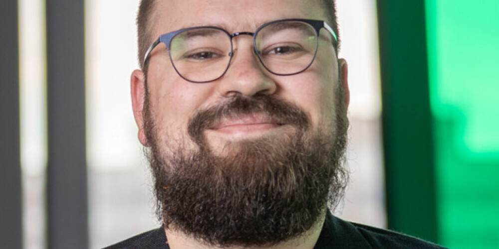 David Duff, a bearded person wearing glasses, a maroon shirt, and a dark blazer, smiles at the camera. The background features large windows with natural light and a green-tinted area.
