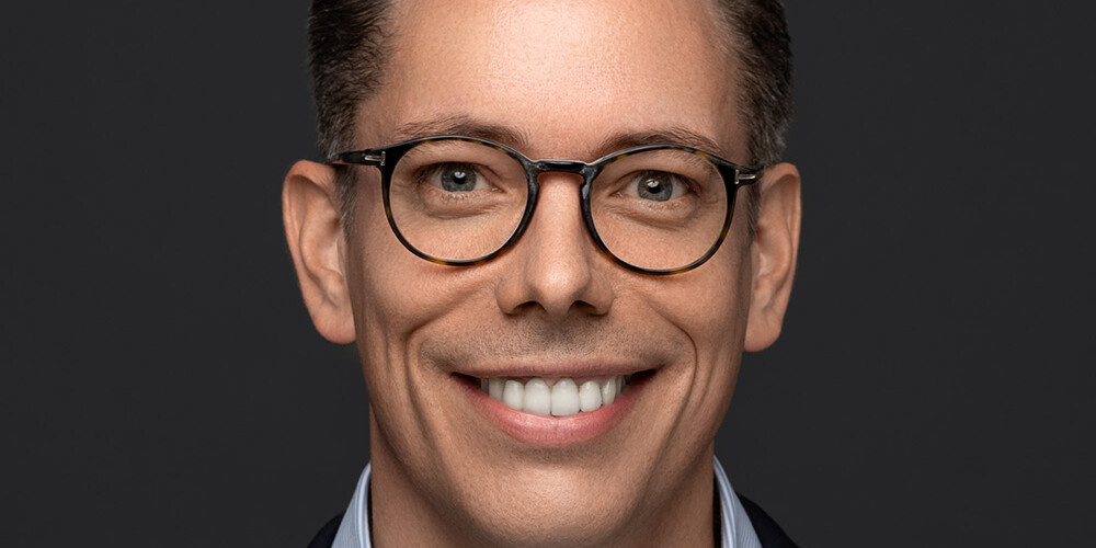David Duff, a man with short brown hair, wearing round glasses, a blue suit jacket, and a light blue striped shirt, smiles in front of a dark gray background.