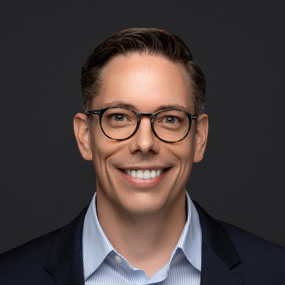 David Duff, a man with short brown hair, wearing round glasses, a blue suit jacket, and a light blue striped shirt, smiles in front of a dark gray background.