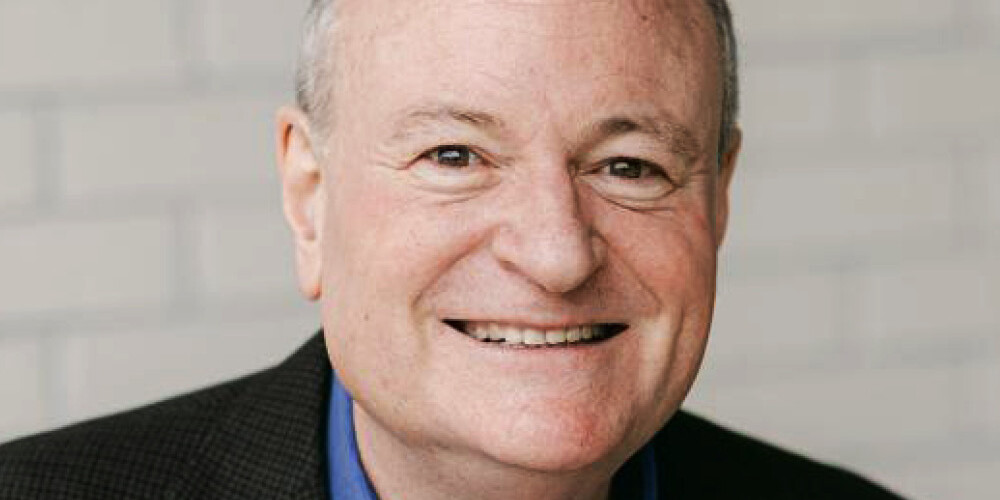 David Duff, a middle-aged man with short gray hair, smiles at the camera. He is wearing a dark blazer over a blue collared shirt and is posed in front of a light-colored brick wall.