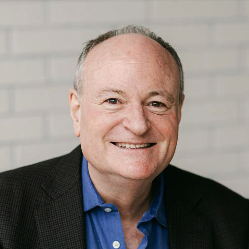 David Duff, a middle-aged man with short gray hair, smiles at the camera. He is wearing a dark blazer over a blue collared shirt and is posed in front of a light-colored brick wall.