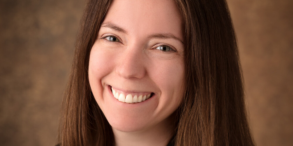 A woman with long straight brown hair is smiling at the camera. She is wearing a black blazer over a light top, posed in front of a brown textured background, reminiscent of the portrait style often favored by David Duff.