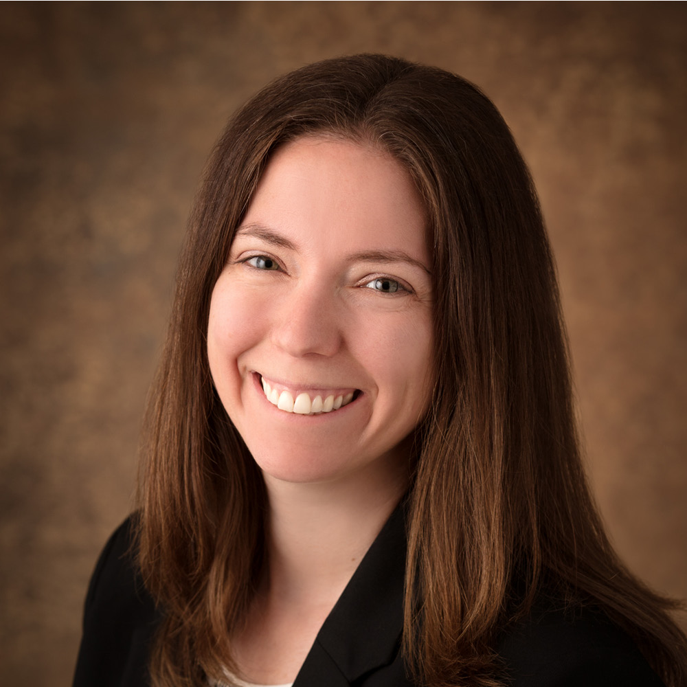 A woman with long straight brown hair is smiling at the camera. She is wearing a black blazer over a light top, posed in front of a brown textured background, reminiscent of the portrait style often favored by David Duff.