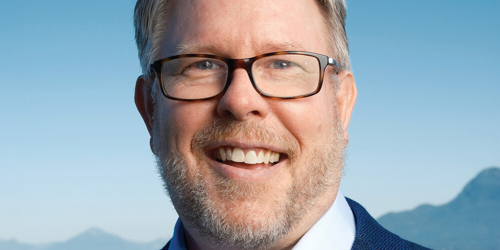 David Duff, a smiling man with short gray hair, glasses, and a beard, wears a blue blazer and light blue shirt while standing outdoors with mountains and a blue sky in the background.