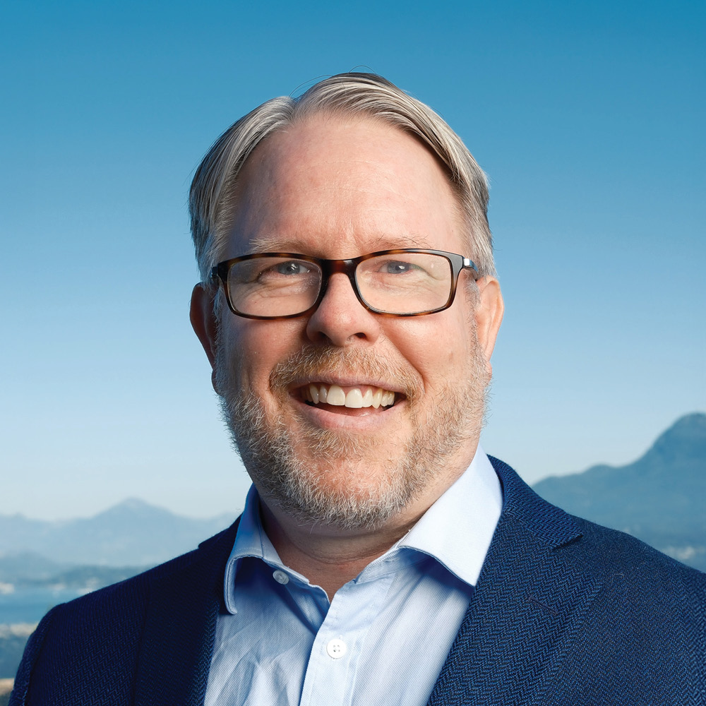 David Duff, a smiling man with short gray hair, glasses, and a beard, wears a blue blazer and light blue shirt while standing outdoors with mountains and a blue sky in the background.