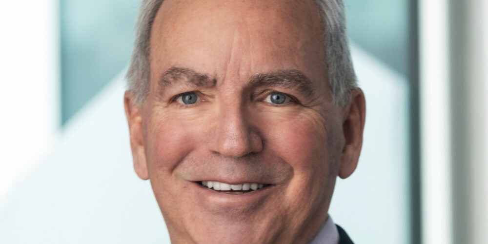 A smiling older man, David Duff, with gray hair, wearing a dark suit, lavender shirt, and purple tie, stands in front of a blurred background featuring large windows and geometric shapes.