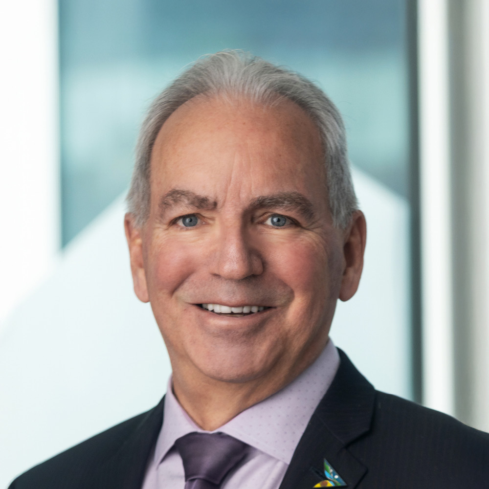A smiling older man, David Duff, with gray hair, wearing a dark suit, lavender shirt, and purple tie, stands in front of a blurred background featuring large windows and geometric shapes.