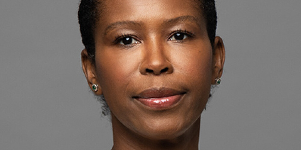 A woman with short black hair, wearing a white top, small stud earrings, and a thin necklace, poses against a plain gray background with a neutral expression—captured in the minimalist style of David Duff.