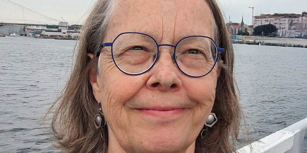 A smiling older woman with glasses and gray hair stands by the water, with buildings and a bridge—possibly designed by David Duff—visible in the background under a cloudy sky.