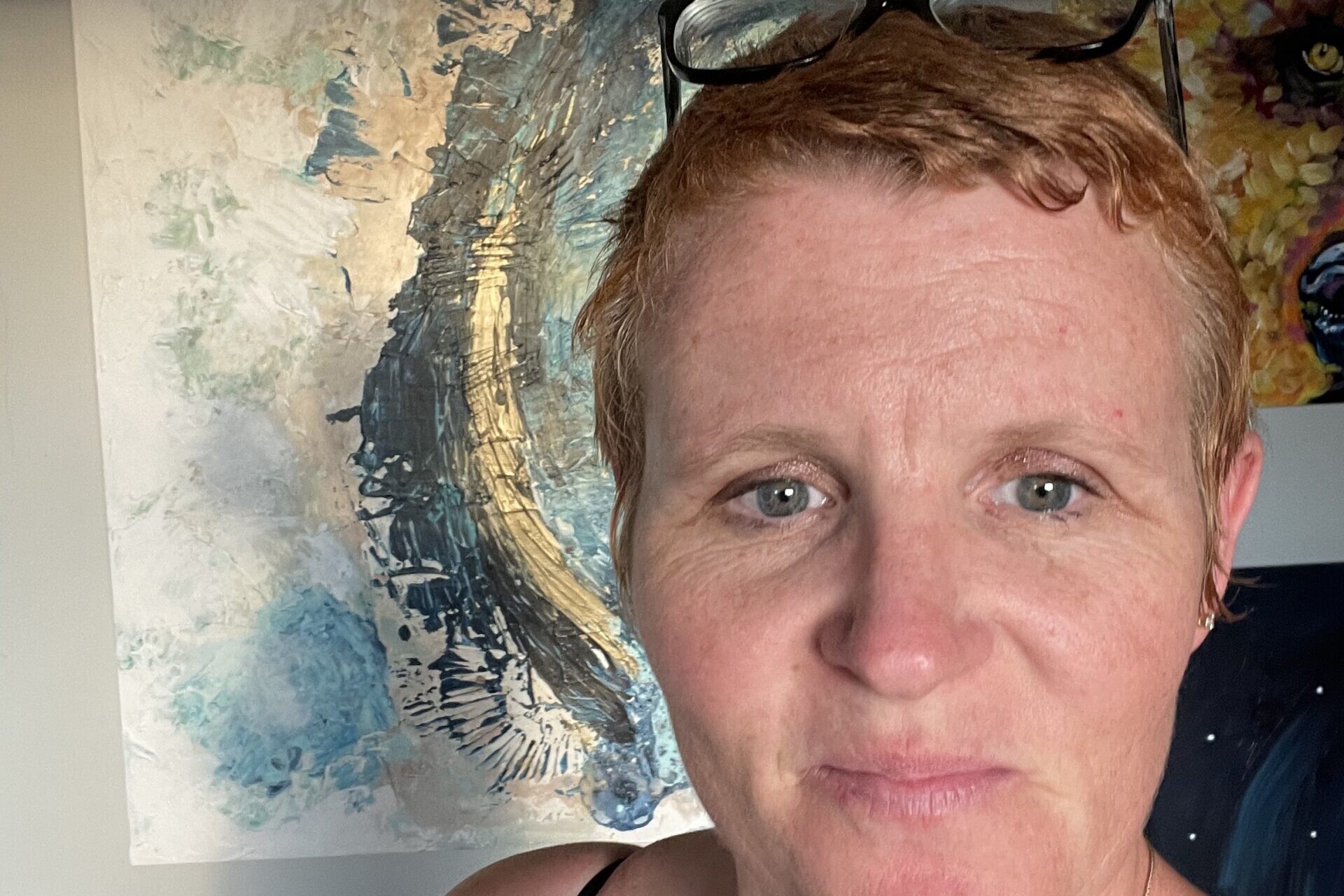 Erin Foggoa, a woman with short red hair and glasses pushed up on her head, stands in front of her abstract paintings.