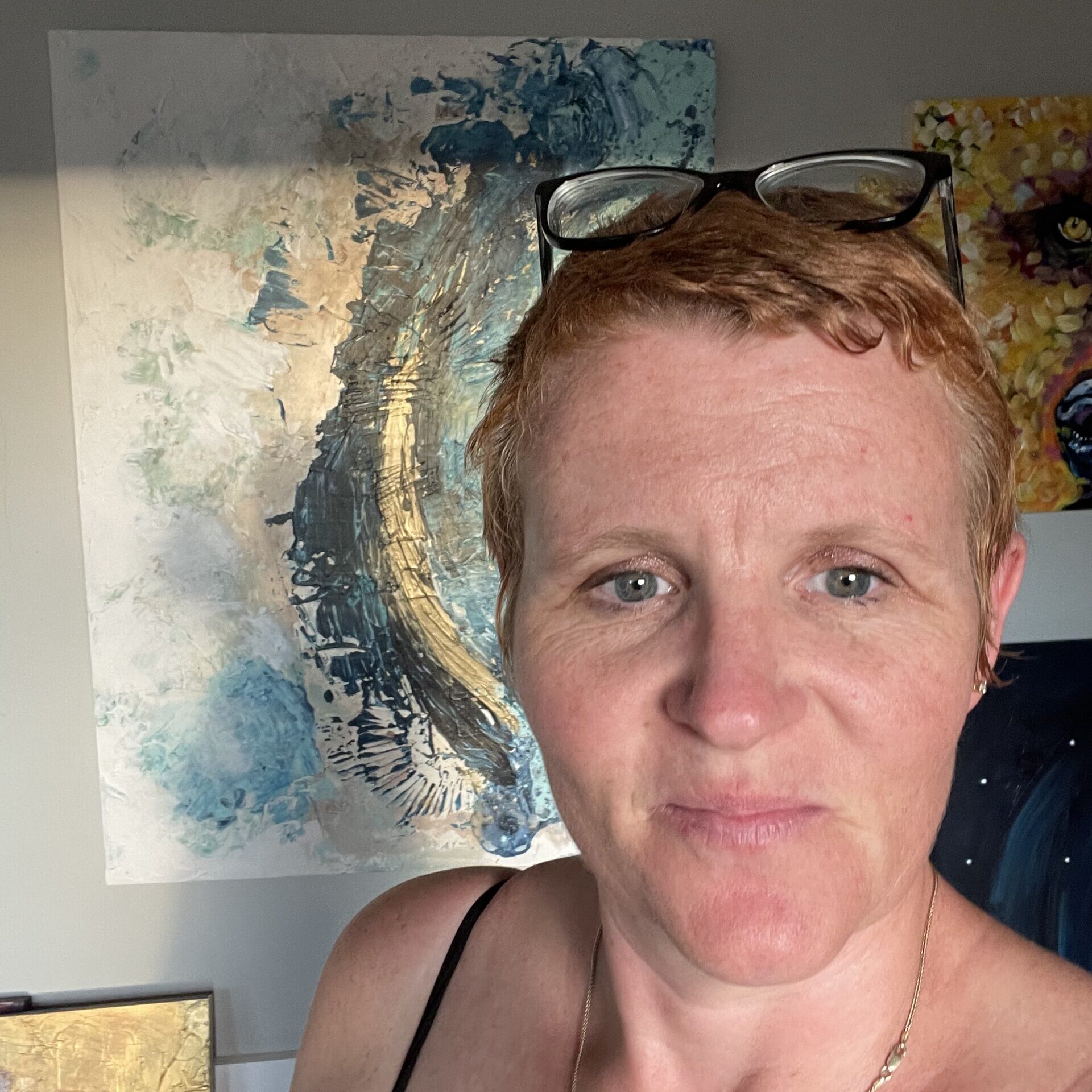 Erin Foggoa, a woman with short red hair and glasses pushed up on her head, stands in front of her abstract paintings.